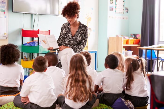 Female Teacher Reading Story To Group Of Elementary Pupils Wearing Uniform In School Classroom