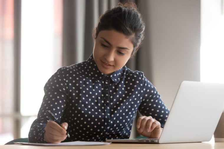 Young indian business woman student working studying online with laptop making notes sit at office desk, focused hindu female professional preparing report doing paperwork writing essay at workplace