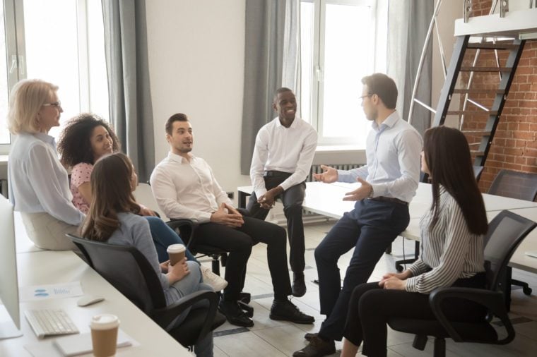 Multiethnic work group talk during casual office meeting, discuss business ideas sharing thoughts, smiling diverse colleagues or employees speak negotiating at informal briefing at workplace