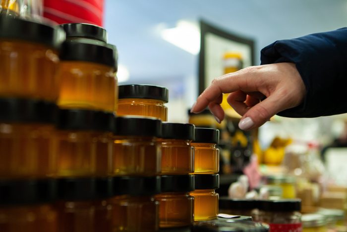 Seasonal products sold at Xmas market. A woman's hand is seen close-up, picking a jar of homemade honey from a shelf at a festive farmer's exposition, with copy-space to the left.