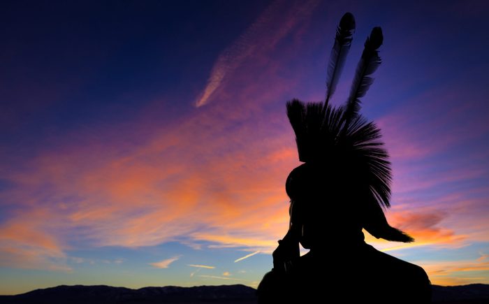 Native American Indian wearing head dress in silhouette against sunset background.