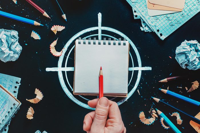 a hand holds a pencil over a small notebook that sits on top of chalk drawn crosshairs. scattered around is other pencils, shavings, and scraps of paper