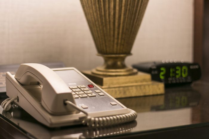 Telephone at hotel room sitting on bedside table along with the table lamp and alarm clock.