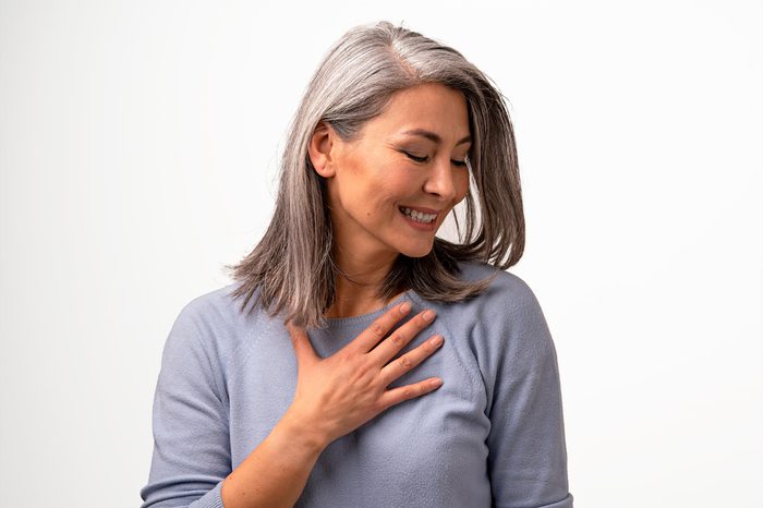 Pleased Middle-Aged Asian Woman Touches Her Chest Thanking. Smiling Woman Touching Her Chest With Hand And Looking Aside While Being Pleased. Portrait. Gratitude Concept