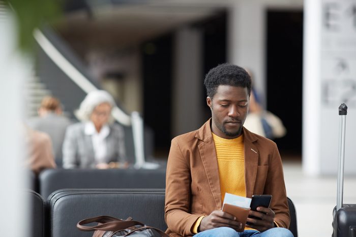Serious pensive young black man with beard sitting in airport and communicating via online app on phone