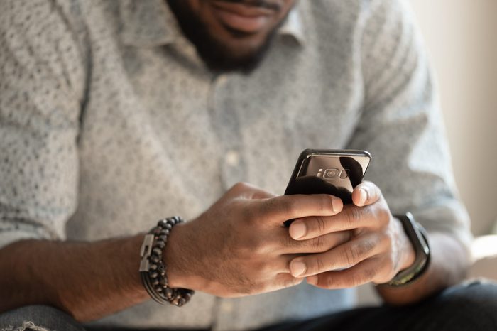 Close up young african american man sitting on couch, holding smartphone in hands, typing message, posting in social media, chatting with friends or relatives, playing online game, doing purchases.