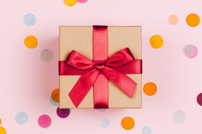 Present box with red bow on pastel pink background with multicolored confetti. Flat lay style.