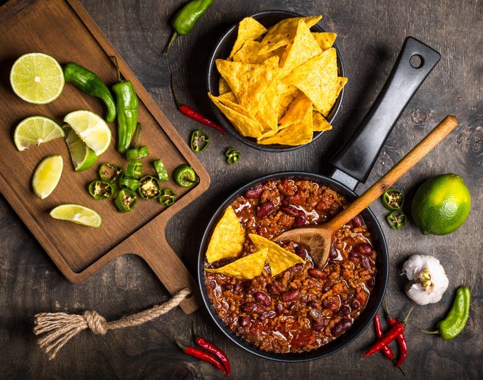 Chili con carne in frying pan on dark wooden background. Ingredients for making Chili con carne. Top view. Chili with meat, nachos, lime, hot pepper. Mexican/Texas traditional dish Chili con carne