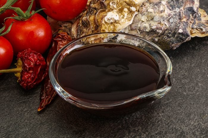oyster sauce in a small glass bowl near oyster shell and tomatoes on dark slate background