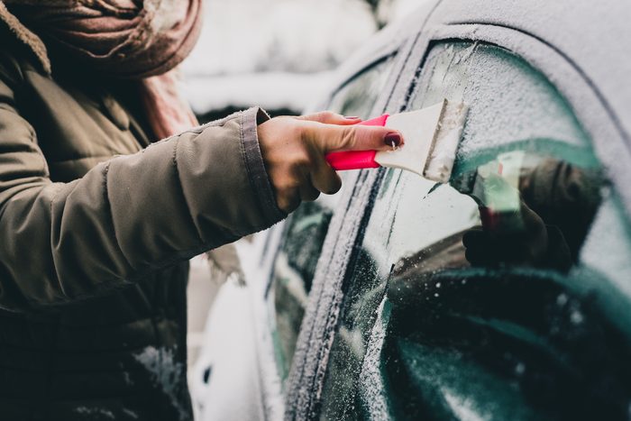 A woman is cleaning an icy window on a car with ice scraper. Focus on the ice scraper. Cold snowy and frosty morning.