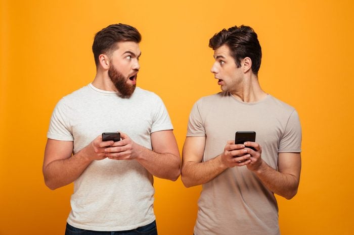 Portrait of a two shocked young men holding mobile phones and looking at each other isolated over yellow background