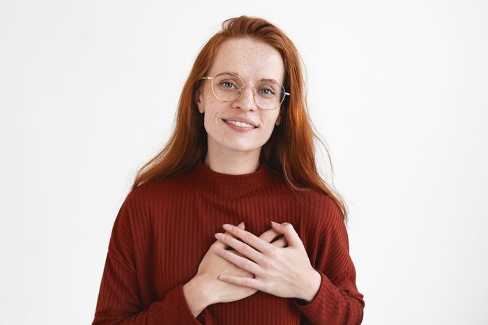 Adorable ginger girl in eyewear keeping both hands on her chest and smiling joyfully, looking with love and gratitude at camera, expressing thankfulness to friend for help and advice, being grateful