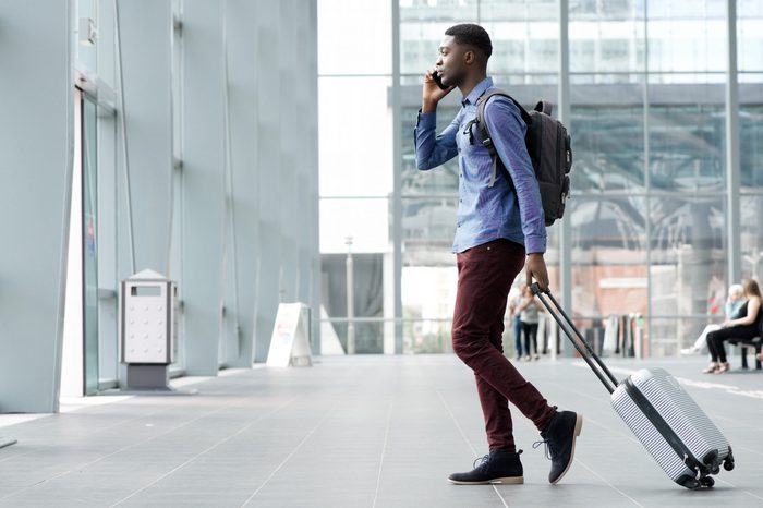 Full body side portrait of young african man traveling with suitcase and cellphone at airport 