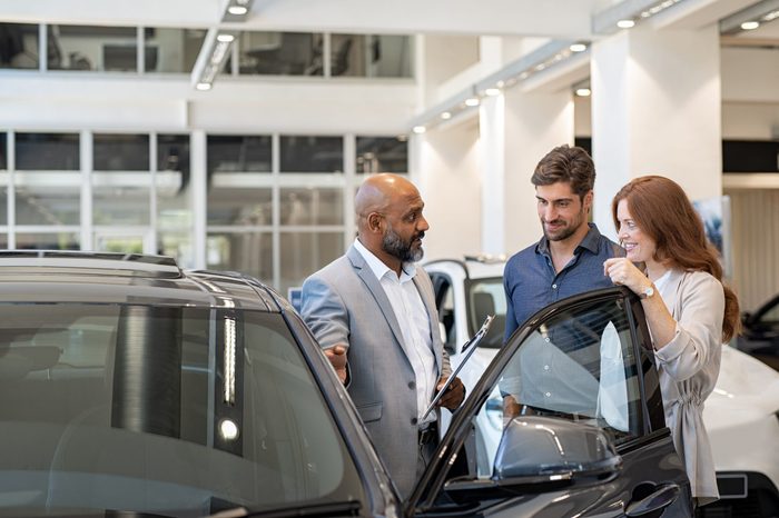 Beautiful couple smiling while examining a new car in a motor show with salesman. Mature salesman showing new luxury car to man and woman in showroom. Happy couple with cardealer showing new features.