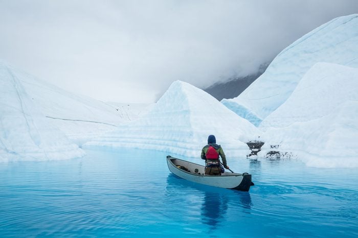 Man paddles a full size inflatable canoe across a deep blue glacier lake carved by water of the melting Matanuska Glacier in Alaska.
