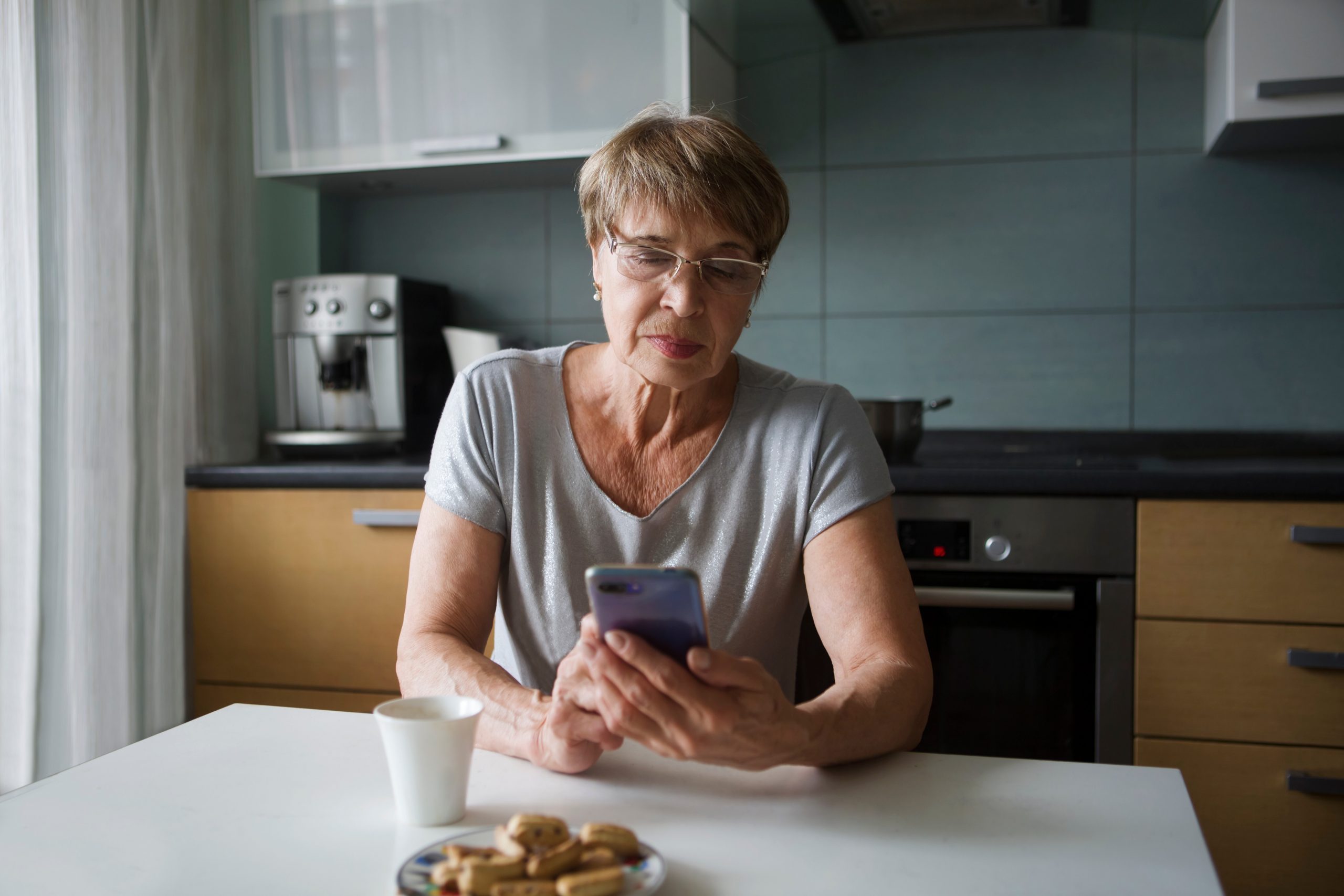 Portret happy senior woman looking at screen of mobile phone while in kitchen 