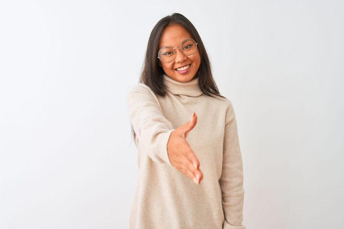 Young chinese woman wearing turtleneck sweater and glasses over isolated white background smiling friendly offering handshake as greeting and welcoming. Successful business.