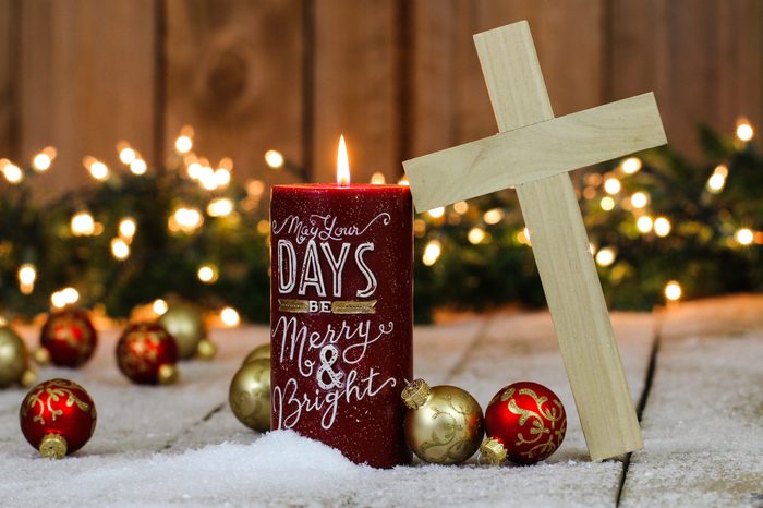 Christmas background with red candle, wood cross, red and gold ornaments and string of holiday lights with green garland border in snow; Christmas religious background
