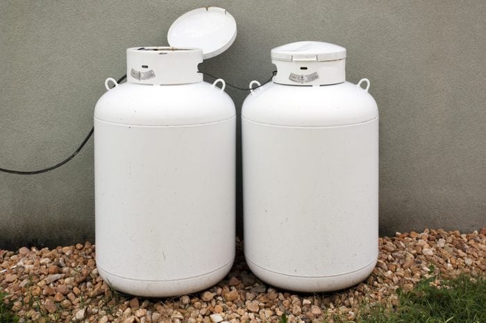 Two home use propane tanks stored outside against building wall with a gravel foreground. Horizontal.