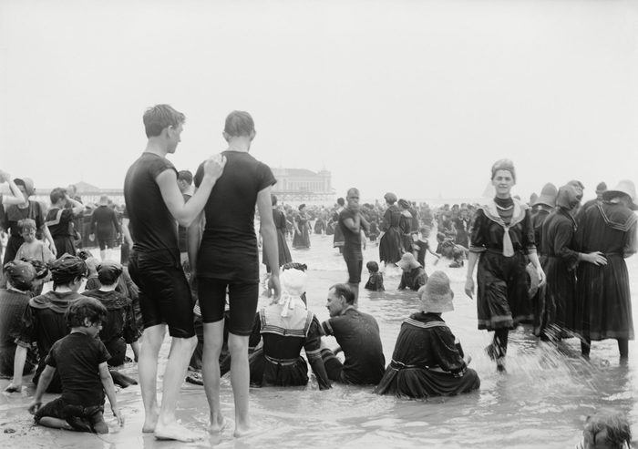 Men, women, and children enjoying the surf at Atlantic City, New Jersey, c. 1890-1900. Most women wear a middy style swimming dress with fill long sleeves and stockings. The men wear short sleeves t-shirts with long shorts
