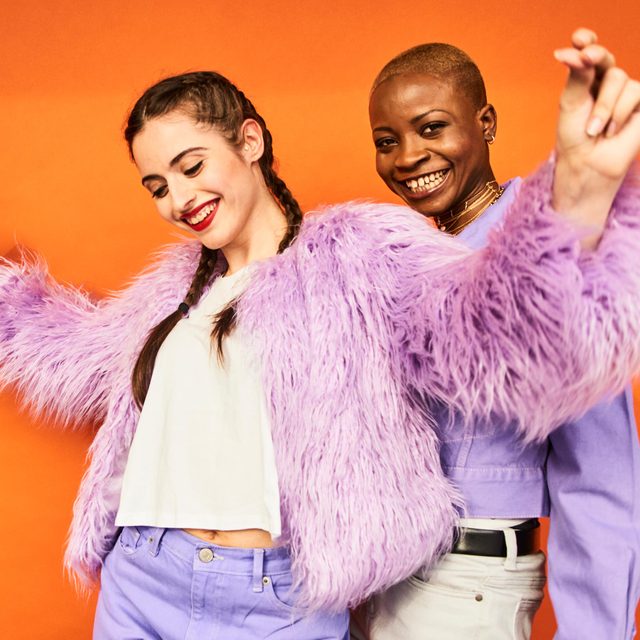 Two young women in purple clothes dancing in front of an orange wall
