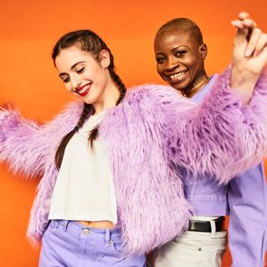 Two young women as best friends dancing together in purple clothes in front of an orange wall