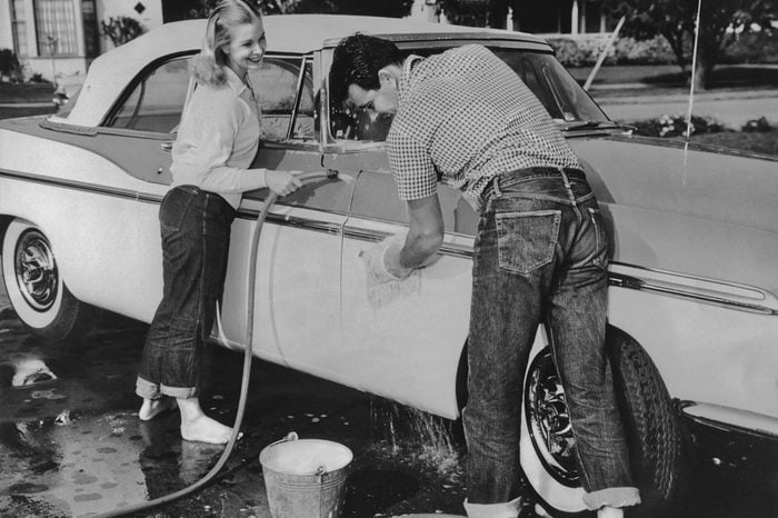 A young man and woman, wearing rolled up jeans, smile while washing the car in a driveway in the 1950's
