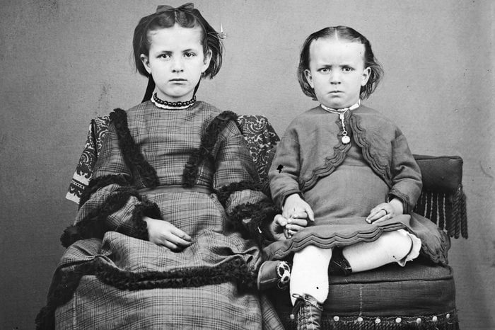 Portrait of two unidentified children holding hands while posed on homemade fancy chairs, Wisconsin, 1869. The girl is dressed in a hand-me-down dress cut from a heavy wool woman's dress while the boy on the right wears an old-fashioned infant's dress which has become too small for him