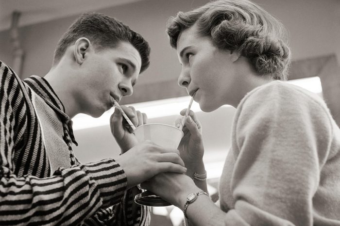 1950s TEENAGE COUPLE SHARING ICE CREAM SODA AT LUNCH COUNTER TWO STRAWS ONE DRINK