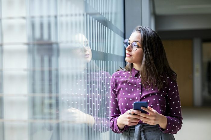 Shot of a young female business professional with a cell phone standing by a glass wall in office and looking away