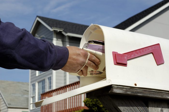 Mailman's arm inserting a bundle of mail into a mailbox. Partially obscured suburban home in background. Horizontal orientation.