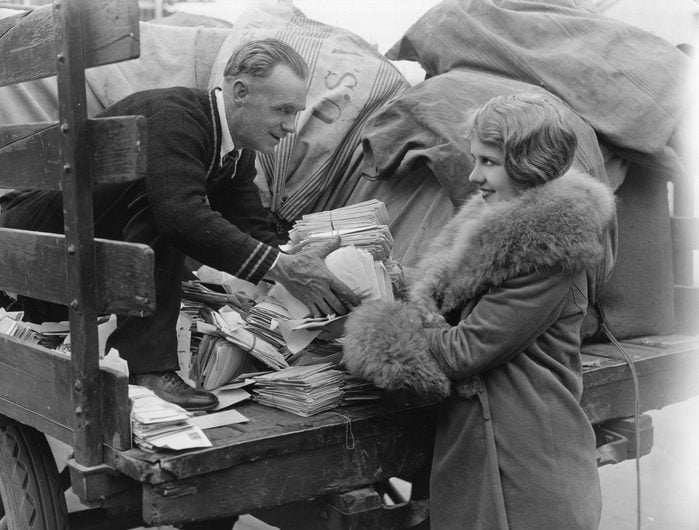 1925: A postman employed by the US mail hands a woman a pile of letters from the back of his lorry.