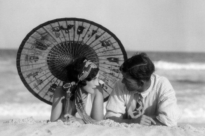 1920s ROMANTIC COUPLE MAN WOMAN LOOKING AT ONE ANOTHER LYING FACE TO FACE UNDER ORIENTAL PARASOL ON SANDY BEACH