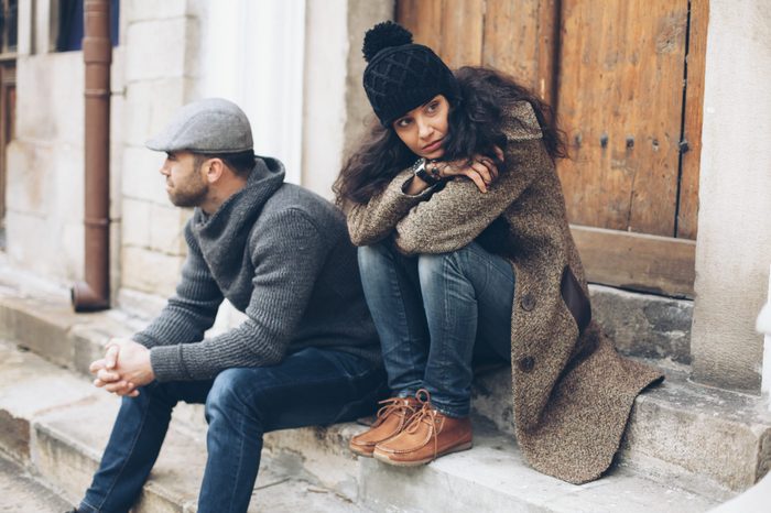 Young couple having relationship difficulties. Man and woman sitting on stairs in front of a closed wooden door. Wears warm clothes - jeans, sweater, coat, hat and scarf. Both looking in different directions. On background building facade, wooden front door, rainwater gutter and windows.