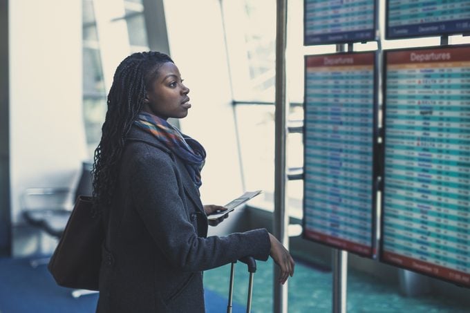 Young African American woman at airport checks the arrivals and departures board with suitcase and passport in hand