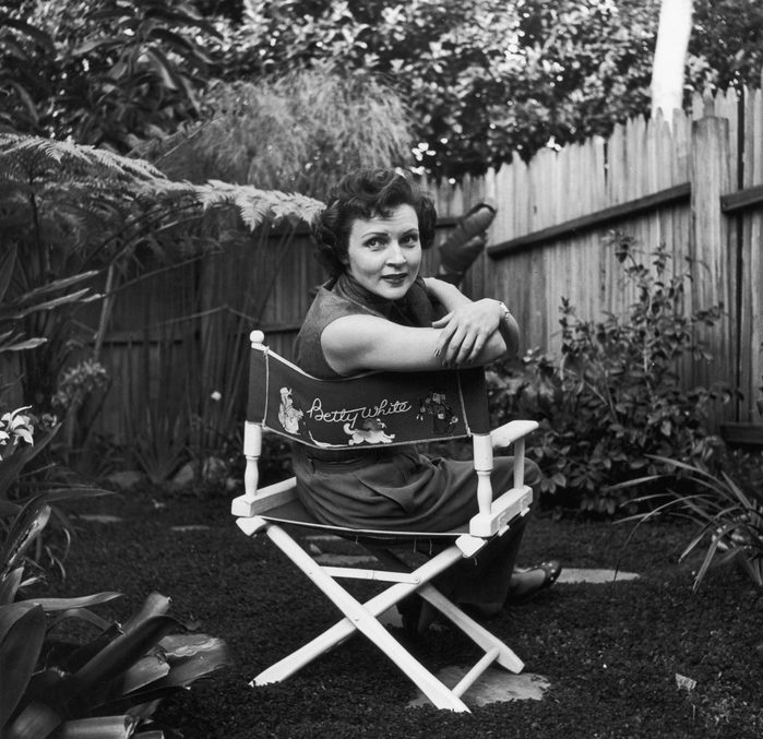 circa 1956: American actor Betty White sits in a canvas chair with her name written on the back, looking over her shoulder in a backyard garden.
