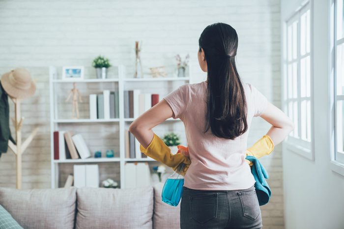 back view of asian housekeeper looking at the clean living room after she tidied up. young wife finished house chores putting hands in waist watching the bookshelf beside the sunlight window.