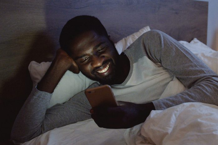 Young handsome African American man lying in the bed in his pajama and chatting with somebody cheerfully on the smartphone. At night.