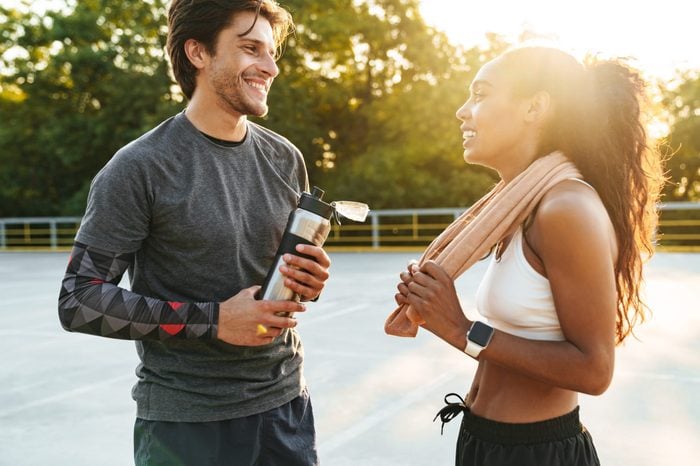 Image of a cheerful pleased young strong sports woman and man holding towel and water talking with each other outdoors.