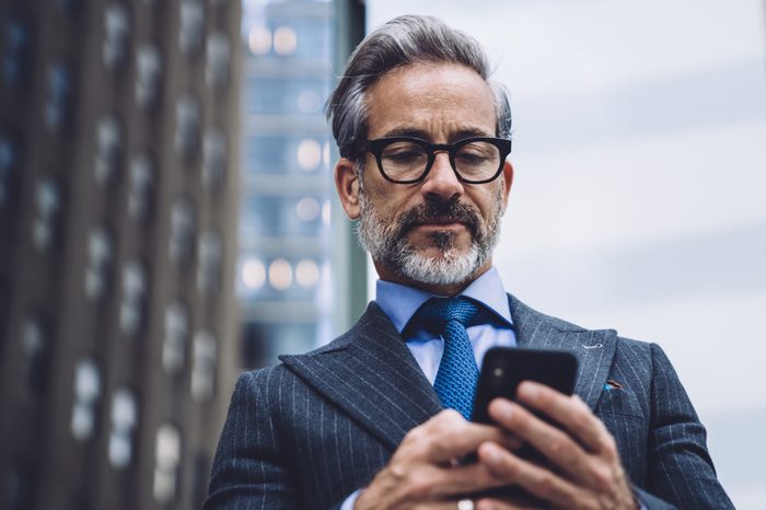 Low angle of elegant grey-haired mature male manager in expensive suit and glasses browsing smartphone on blurred background with New York skyscrapers