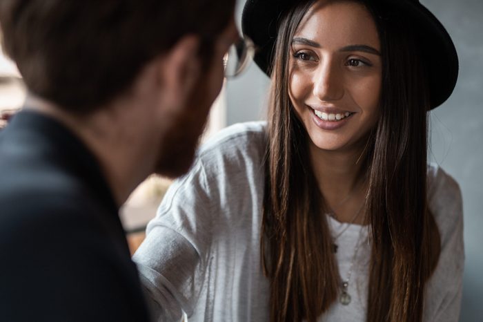 Cheerful young woman in hat smiling and talking with anonymous man during romantic date in cafe