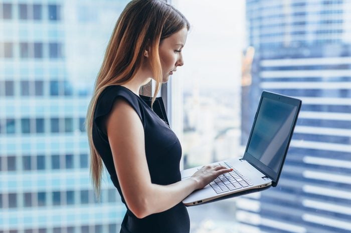 Portrait of young woman working holding laptop standing against panoramic window with city view