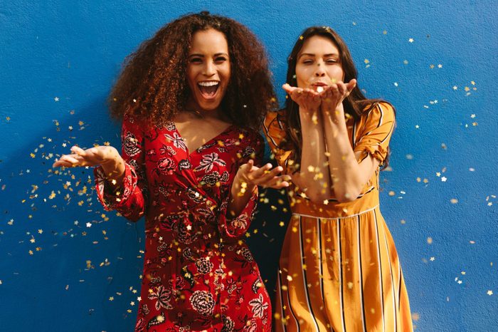 Female friends on galentines day blowing glitter off their hands standing against a blue wall.