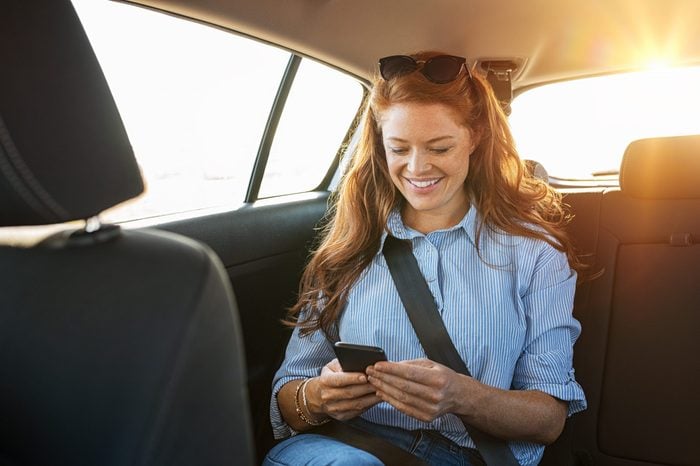 Smiling casual woman sitting in back seat using mobile phone. Cheerful young woman reading messages in smartphone while sitting in a taxi. Attractive girl with red hair wearing blue shirt in car using cellphone.
