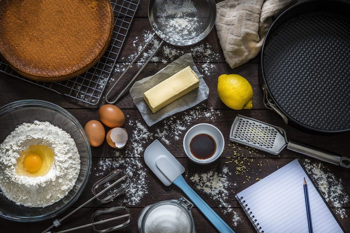 Top view of various ingredients and utensils for baking cakes or cooking desserts like flour, eggs, sugar, butter, a spatula, a sieve, a baking mold and a grater with lemon zest. At the lower right corner is an opened cookbook with copy space ideal for a text, a logo or a recipe. All the objects are on a rustic wooden table. Low key DSLR photo taken with Canon EOS 6D Mark II and Canon EF 24-105 mm f/4L