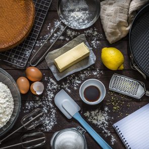 Top view of various ingredients and utensils for baking cakes or cooking desserts like flour, eggs, sugar, butter, a spatula, a sieve, a baking mold and a grater with lemon zest. At the lower right corner is an opened cookbook with copy space ideal for a text, a logo or a recipe. All the objects are on a rustic wooden table. Low key DSLR photo taken with Canon EOS 6D Mark II and Canon EF 24-105 mm f/4L