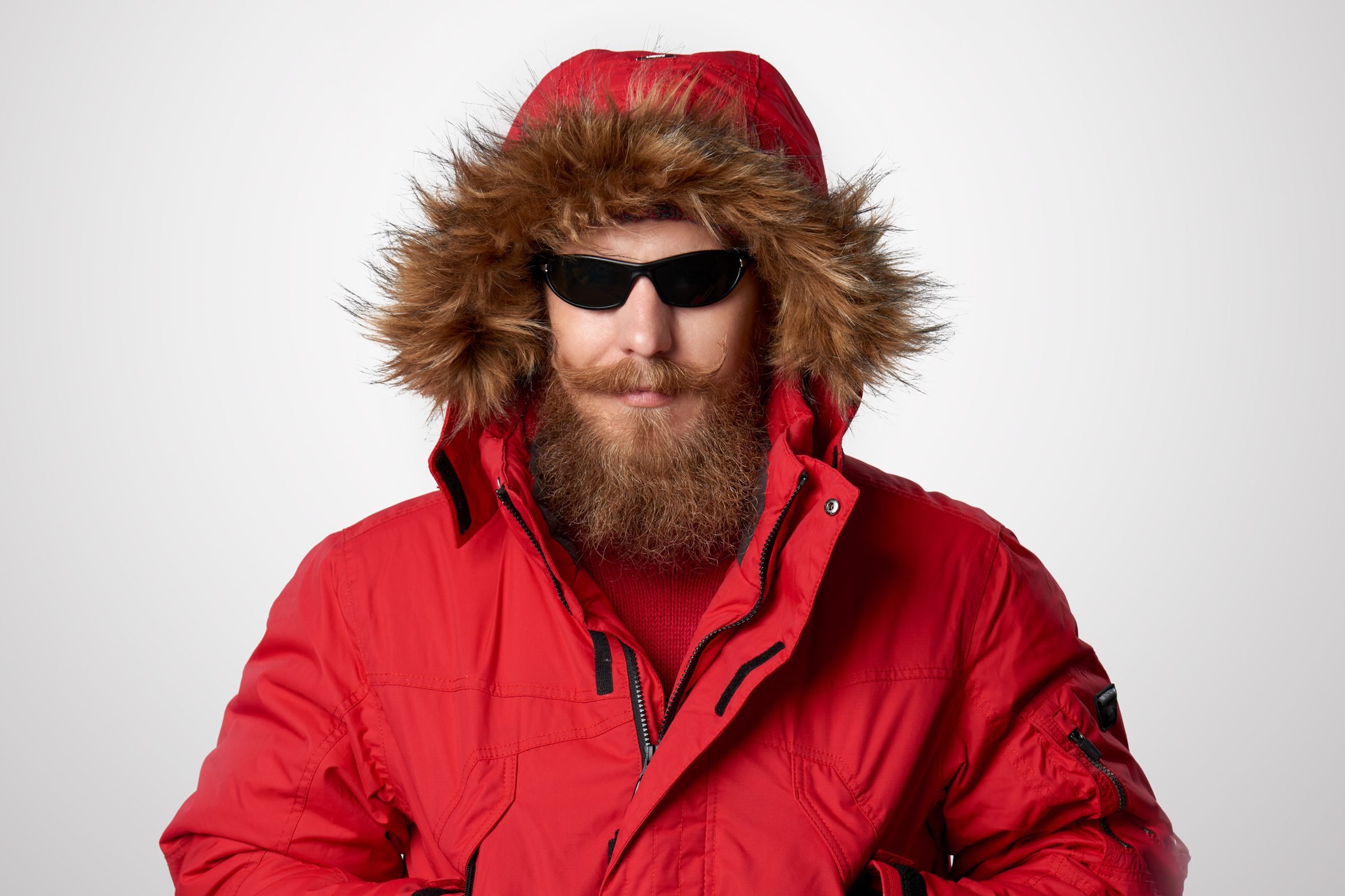 man with a mustache and beard wears sunglasses and a large red winter coat with a fur hood that matches his facial hair