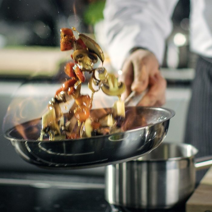 Professional Chef Cooks Flambe Style. He Prepares Dish in a Pan with Open Flames. He Works in a Modern Kitchen with Different Ingredients Lying Around.