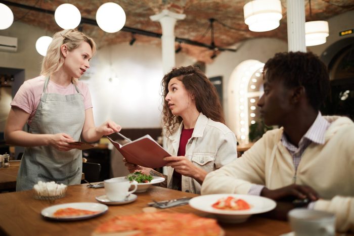 Disgruntled curly-haired young female restaurant guest sitting at table with boyfriend and reading menu while talking to waitress giving advice to her in modern loft cafe