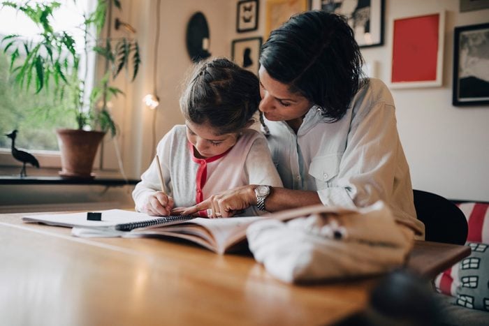 Mother assisting daughter in writing homework on table while sitting against wall at home